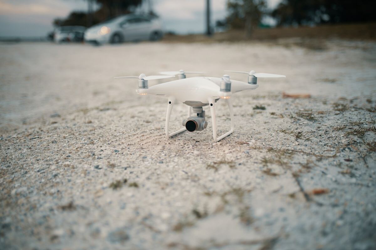 Drone blanc posé sur un sol sablonneux avec des voitures en arrière-plan.
