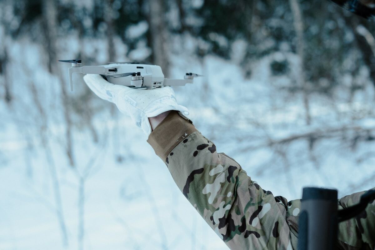 Drone blanc tenu par une personne en tenue de camouflage dans un paysage enneigé.